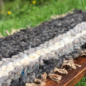 May include: Handmade woven rug with a textured, striped design. The rug features alternating bands of grey, off-white, and black wool. The edges are finished with a fringe detail. The rug is placed on a wooden surface.