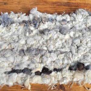 May include: A rectangular, hand-woven rug with a textured, fluffy appearance. The rug features a mix of white, gray, and brown wool fibers, creating a natural, rustic aesthetic. The fibers are loosely woven, giving it a soft, inviting look.