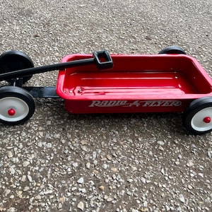 May include: A classic red metal wagon with a black handle and four black wheels featuring white rims and red center caps. The side of the wagon body displays the white text Radio Flyer. The wagon has a rectangular bed with rounded corners.