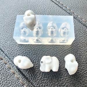 May include: A silicone mold with four cavities shaped like elephant heads. There are four white ceramic elephant charms, each with a hole for stringing.