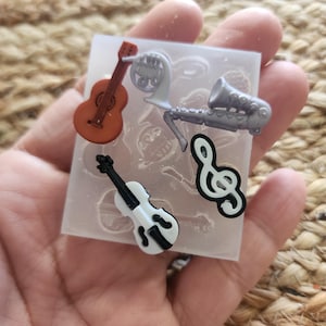 May include: A clear silicone mold with miniature musical instruments. The mold includes a brown guitar, a silver French horn, a silver saxophone, a black and white violin, and a black and white treble clef.