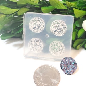 Op de afbeelding: Een doorzichtige siliconen mal met vier cirkelvormige holtes gevuld met een sprankelend, iriserend materiaal. Een afgewerkt ovaal stuk en een kwart dollar munt zijn ook zichtbaar, wat wijst op een knutselproject.