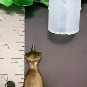 May include: A small, bronze-colored dress form figurine, approximately 1.5 inches tall, next to a clear, cylindrical mold. The dress form is detailed with a classic silhouette. A ruler is in the foreground.