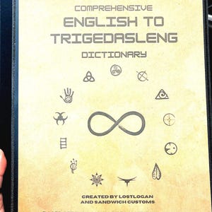 May include: The cover of a book titled "The Unofficial Comprehensive English to Trigedasleng Dictionary". The cover is beige with black text and features a variety of symbols, including an infinity symbol, a triangle, a circle with a cross, and a hand with a spiral.