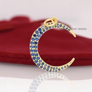 May include: A gold crescent moon charm with a blue sapphire stone design. The charm is on a red velvet surface.