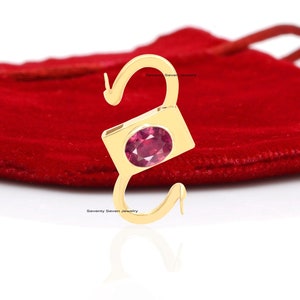 May include: A gold charm with a pink gemstone in the center. The charm is shaped like a sideways 'S' and is on a red velvet background.