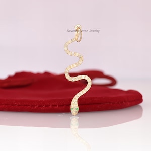 May include: A gold snake charm with a green stone for the eyes, set with clear stones. The charm is hanging from a red velvet bag.