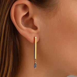 Può includere: Orecchini color oro con un design a barra verticale. Ogni orecchino presenta una barra rettangolare dorata con una piccola gemma blu a forma di goccia che pende dal basso. Gli orecchini sono mostrati su un orecchio.