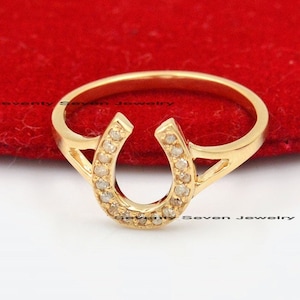 Könnte beinhalten: Ein goldener Hufeisenring mit kleinen, klaren Steinen, die in die Hufeisenform eingefasst sind. Der Ring liegt auf einem roten Samt-Hintergrund.