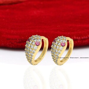 May include: A pair of gold hoop earrings with a small red stone and white cubic zirconia stones. The earrings are on a white background with a red velvet pouch.