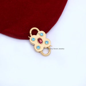 Op de afbeelding: Gouden, bloemvormige sluiting met een rood en blauw edelsteenontwerp. Seventy Seven Jewelry.