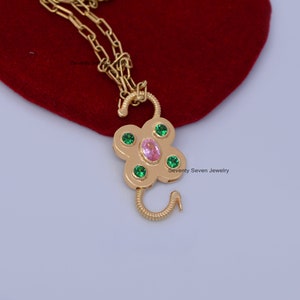 Op de afbeelding: Gouden ketting met een bloemvormige hanger met roze en groene edelstenen. De hanger is in de vorm van een klavertjevier met een haaksluiting. De ketting ligt op een rode fluwelen achtergrond. "Seventy Seven Jewelry" staat op de achtergrond.
