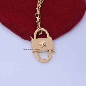 Op de afbeelding: Gouden hangslotbedel met een sterontwerp. De bedel zit aan een gouden ketting en hangt aan een rood fluwelen oppervlak. "Seventy Seven Jewelry" staat op het oppervlak.