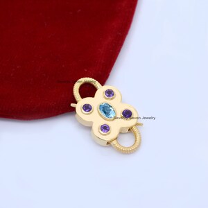Op de afbeelding: Gouden metalen sluiting met een bloemvormig ontwerp. De sluiting heeft een blauwe ovale edelsteen omringd door vijf kleinere paarse edelstenen.
