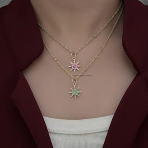 May include: A gold chain necklace with two star-shaped pendants. The top pendant is pink and the bottom pendant is green.