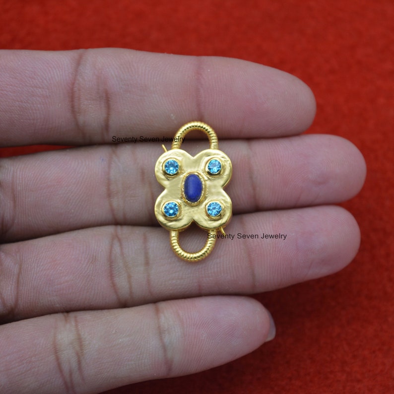 Op de afbeelding: Gouden metalen sluiting met een blauwe steen en vier kleinere blauwe stenen. De sluiting is in de vorm van een klavertjevier. "Seventy Seven Jewelry" staat op de sluiting.