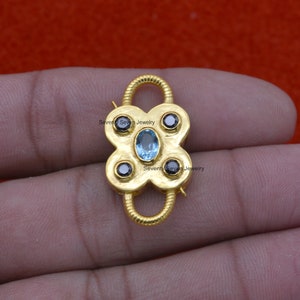 Op de afbeelding: Gouden metalen sluiting met een blauwe edelsteen en vier zwarte edelstenen. "Seventy Seven Jewelry" staat op de sluiting.