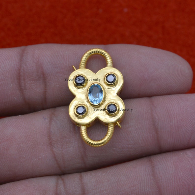 Op de afbeelding: Gouden metalen sluiting met een blauwe edelsteen en vier zwarte edelstenen. "Seventy Seven Jewelry" staat op de sluiting.