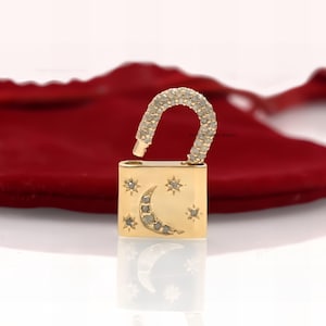 May include: A gold padlock charm featuring a crescent moon and stars design, set with clear diamonds. The padlock is open and the charm is displayed on a red velvet background.