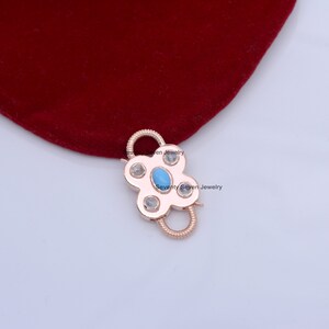 Op de afbeelding: Roze gouden metalen sluiting met een turquoise en wit edelsteen ontwerp. De sluiting is gevormd als een bloem met vier bloemblaadjes.  Seventy Seven Jewelry is op de sluiting gedrukt.