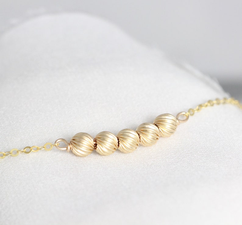 May include: A gold chain necklace with a row of five gold beads. The beads have a textured, ribbed surface.