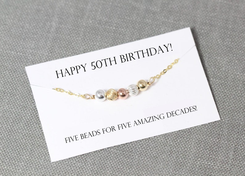 May include: A white card with the text "HAPPY 50TH BIRTHDAY!" and "FIVE BEADS FOR FIVE AMAZING DECADES!" A gold chain with five beads in different colors is on the card. The beads are silver, gold, rose gold, and a textured gold.