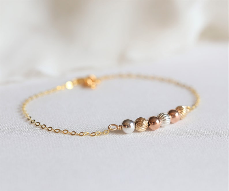 May include: A gold chain bracelet with a row of five beads in different shades of gold, silver, and rose gold.