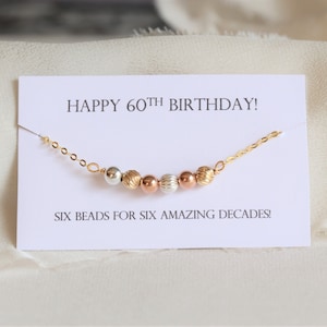 May include: A gold chain necklace with six beads in different colors and textures. The card reads "Happy 60th Birthday! Six beads for six amazing decades!"