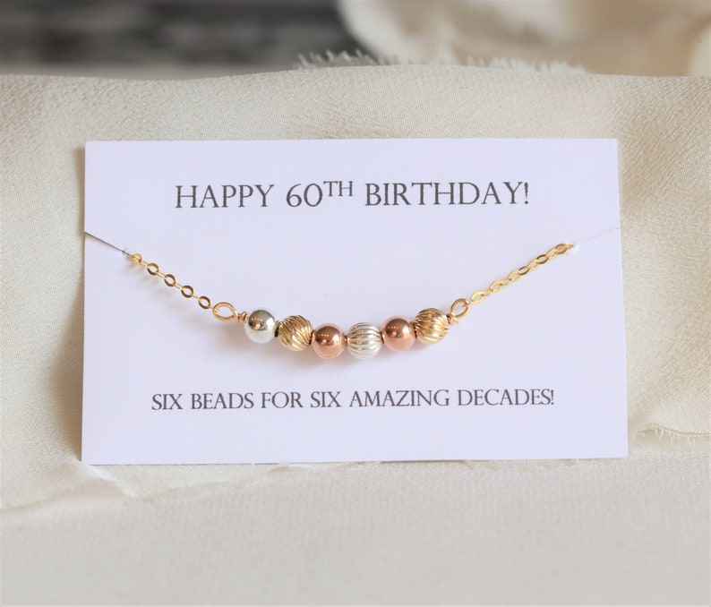 May include: A gold chain necklace with six beads in different colors and textures. The card reads "Happy 60th Birthday! Six beads for six amazing decades!"
