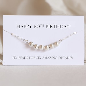May include: A white card with the text "HAPPY 60TH BIRTHDAY!" and "SIX BEADS FOR SIX AMAZING DECADES!"  A silver chain with six silver beads is displayed on the card.