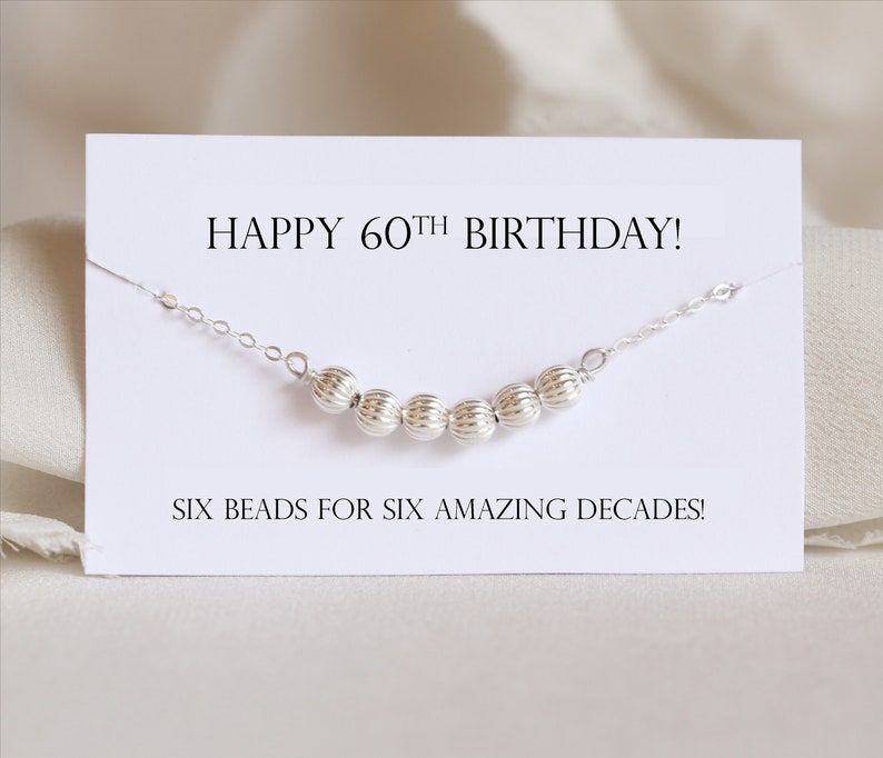 May include: A white card with the text "HAPPY 60TH BIRTHDAY!" and "SIX BEADS FOR SIX AMAZING DECADES!"  A silver chain with six silver beads is displayed on the card.