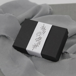 May include: A black gift box with a white label featuring a floral design. The label has a simple line drawing of a flower with leaves.