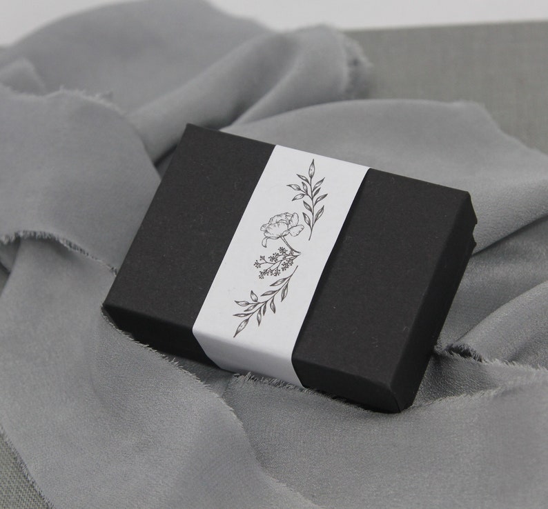 May include: A black gift box with a white label featuring a floral design. The label has a simple line drawing of a flower with leaves.