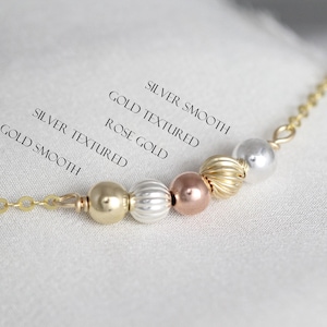 May include: A close-up of a necklace with beads in different colors and textures. The beads are labeled "Gold Smooth", "Gold Textured", "Rose Gold", "Silver Textured", and "Silver Smooth".
