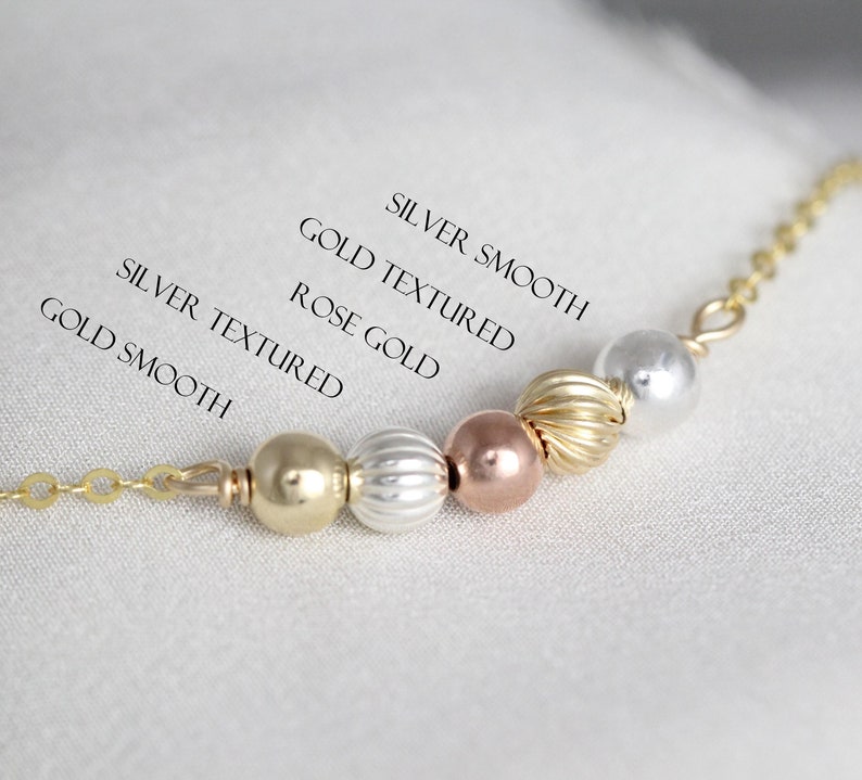 May include: A close-up of a necklace with beads in different colors and textures. The beads are labeled "Gold Smooth", "Gold Textured", "Rose Gold", "Silver Textured", and "Silver Smooth".