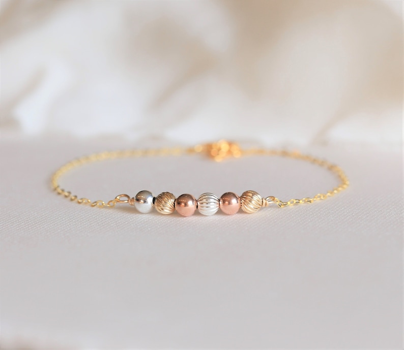 May include: A gold chain bracelet with five beads in different colors: silver, gold, rose gold, gold, and silver. The beads are round and have a textured surface.
