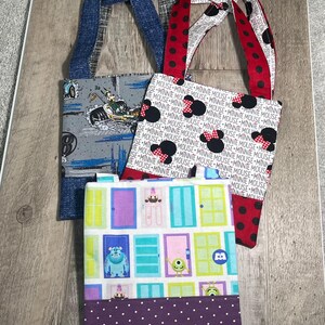 May include: Three fabric tote bags with different patterns. One has a gray background with a cartoon character on a motorcycle. Another has a white background with Minnie Mouse graphics. The third has a purple polka dot bottom and a door design.