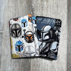 May include: Two fabric wallets featuring designs from The Mandalorian. One wallet has a white background with colorful Mandalorian helmet graphics. The other wallet has a black background with a Mandalorian character illustration and text.
