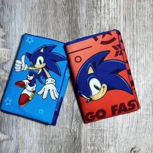 May include: Two rectangular fabric items featuring Sonic the Hedgehog. The left item is blue with a full-body graphic of Sonic giving a thumbs up and small star outlines. The right item is orange with a close-up of Sonic and the text GO FAST.