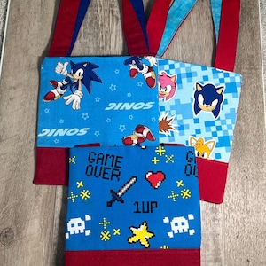 May include: Three blue tote bags with red accents and straps. Each bag features a different design with characters from the Sonic the Hedgehog video game series. One bag has the text "GAME OVER" and pixelated graphics.