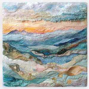 May include: Textile art piece depicting a landscape with layered fabric. The artwork features a sunset with orange and pink hues, blue mountains, and textured clouds. The piece is square and has a quilted appearance.