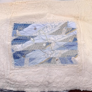 Peut inclure: Un oreiller en crochet blanc avec un motif de patchwork bleu et blanc au centre. Le patchwork est encadré de dentelle blanche.