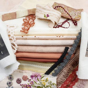 Könnte beinhalten: Ein Stapel weißer und cremefarbener Stoffreste mit Spitzenbesatz, Stickerei und verschiedenen Texturen. Die Stoffreste sind in Schichten angeordnet, wobei die oberste Schicht ein weißer Stoff mit Spitzenbesatz ist. Die unterste Schicht ist ein cremefarbener Stoff mit einem floralen Stickmuster. Die Stoffreste sind von verschiedenen anderen Stoffresten umgeben, darunter ein rot-weißes Stoff mit einem Blumenmuster, ein braunes Stoff mit einem geometrischen Muster und ein schwarzes Stoff mit einem einfarbigen Muster.