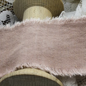 May include: Close-up of a dusty rose-colored linen ribbon with a frayed edge. The ribbon is displayed on a wooden spool, with white lace in the background. The fabric has a woven texture.