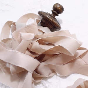 Peut inclure: Un tas de ruban de soie rose pâle aux bords effilochés, posé sur une surface blanche. Une bobine en bois avec un bouton est placée sur le ruban.