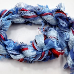 May include: A looped, braided textile in shades of blue, with red and silver accents. The yarn has a fluffy, textured appearance, with strands of varying thicknesses. The craft material is suitable for various DIY projects. The design is looped, creating a circular shape.
