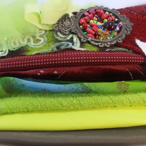 Puede incluir: Una pila de telas y adornos coloridos, incluyendo textiles rojos, verdes y amarillos. Un marco decorativo plateado contiene cuentas y gemas. Se adjuntan fotos de estilo vintage. Un lazo de tul verde lima y otros elementos decorativos son visibles.