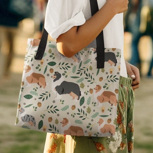 May include: A beige tote bag with a repeating pattern of brown and white dogs, green leaves, and brown acorns. The bag has black straps.