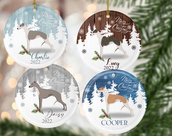 Op de afbeelding: Vier ronde witte kerstballen van keramiek met een winters boslandschap. Elke bal heeft een greyhound-hondenras, een besneeuwd bos en de tekst "Merry Christmas". De ballen zijn gepersonaliseerd met de namen "Charlie", "Lucy", "Daisy" en "Cooper" en het jaar "2022".
