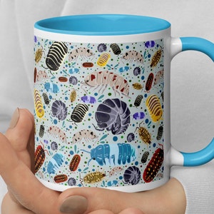 May include: A white ceramic mug with a blue rim and handle. The mug features a colorful pattern of various types of pill bugs, also known as roly-polies, on a light blue background.